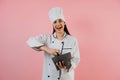 Younh hispanic woman wearing chef hat with ingredients on traditional mortar to cook mexican food on pink color background in Mexi