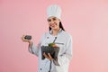 Younh hispanic woman wearing chef hat with ingredients on traditional mortar to cook mexican food on pink color background in Mexi