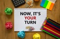 Your turn symbol. White note with words `now it is your turn` on beautiful wooden table, colored paper, colored pencils, paper