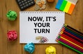 Your turn symbol. White note with words `now it is your turn` on beautiful wooden table, colored paper, colored pencils, paper