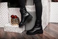 Youth fashionable leather black women`s winter boots. Young woman in jeans in trendy shoes stands in a room near the holiday boxe