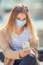 Youth girl sitting outdoors texting on a cellphone wearing protective face mask