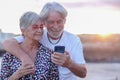 Youthful senior couple embracing in outdoor at sea at sunset using mobile phone. Caucasian retired enjoying relax and happy