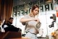 A youthful thin blonde girl,wearing casual cothes,is shown adding milk to the coffee in a cozy coffee shop.