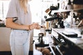 A youthful thin blonde lady,wearing casual clothes,stands nexrt to the coffee machine in a cozy coffee shop.