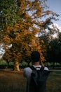 Yping photographer with backbag shootong a long autumn tree in the park under blue sky