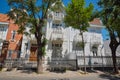 Old white house made of wood, shutters and balconies made of wood. Large deciduous trees grow nearby.