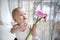 1 little blonde girl with beautiful blue eyes sitting by the window with pink flowers