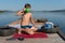 A 11-year-old boy drinks water from a mug while sitting on a wooden pier against the shore and lake on a sunny summer day