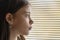An 11-year-old girl looks through the lowered blinds of the window at the street, close-up, selective focus. Concept: waiting for