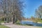 2 ducks cross a beaten path or trail at spring. On the water canal lies an empty mooring pier. A person takes photos of bare trees