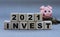 2021 Invest - words on wooden cubes, next to a pig with a slide of coins on a gray background