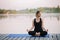 A 36-year-old young Caucasian woman practices yoga in the lotus position outdoors near the river on a wooden pier in the morning.