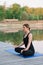 A 36-year-old young Caucasian woman practices yoga in the lotus position outdoors near the river on a wooden pier in the morning.