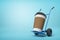 3d close-up rendering of huge paper coffee cup on blue hand truck on light-blue background.