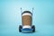 3d close-up rendering of huge paper coffee cup on blue hand truck on light-blue background.