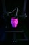 A 3D-FDM-printer standing in dark neon surrounding manufactures a humanoid head from pink plastic.