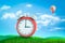 3d rendering of red retro alarm clock showing 3 o`clock standing on green grass under blue sky with hot air balloon in