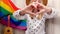 4k Young blonde millennial hippie woman showing heart sign with hands with rainbow LGBTQ flag at home balcony. Peace and