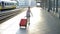 5-6 year old girl is dragging a large red suitcase along the railway platform. Back view.