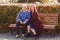 A 50-year-old man in a blue sweater and a woman in a burgundy coat sit on a park bench in the fall