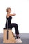 A 50-year-old trainer practices Pilates on an elevator chair, sitting up straight with her arms raised and folde