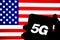 5G letters on a silhouette of a smartphone hold in hand with the flag of the United States on a blurred background screen.