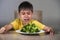 7 or 8 years old upset and disgusted Asian kid sitting on table in front of broccoli plate looking unhappy rejecting the fresh