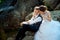 Above outdoor wedding portrait of the charming brude hugging back the happy groom while sitting on the rock.