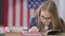 Absorbed smart teen schoolgirl reading out loud sitting on the right at desk in classroom with American flag at