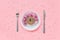 Abstract funny face of woman made donut with eyes and hair from centimeter tape on plate, cutlery on pink background. Fast food,