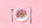 Abstract funny face of woman made donut with eyes and hair from centimeter tape on plate, cutlery on pink background. Fast food,