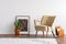 Abstract graphic in wooden frame next to green plant in ceramic pot and elegant beige armchair and orange magazine rack, real