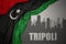 Abstract silhouette of the city with text Tripoli near waving colorful national flag of libya on a gray background