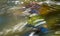 Abstract stream of muddy water flowing over green and brown boulder in wild river bed