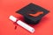 Academic cap with tassel and diploma with ribbon on red surface.