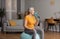 Active aged woman drinking water from sports bottle, sitting on fitness ball in living room interior, copy space