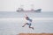 Active jumping kid in shirt and shorts on ocean coast with cargo ship on skyline during spring leisure activity