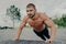 Active muscular man does push up exercise poses shirtless outdoor, exercises in park, has thick beard, stands in lower position.