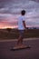 Active teenage boy riding a skateboard on road and admiring scenic landscape during sunset against cloudy sky. Sporty boy riding