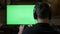 Addictive teenager wearing headphones chilling and playing video game using game controller on green screen TV at home -