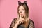 Adolescent in black dress, headband like cat ears, face painting. She showing be quiet sign, posing on pink background. Close up