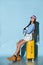 Adolescent in jeans overall skirt, sweatshirt, boots, sun visor cap. She sitting on yellow suitcase, blue background. Full length
