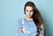 Adolescent in jeans overall, striped sweatshirt. She looking offended, posing with folded hands on blue background. Close up