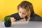 Adorable blonde inquisitive girl with focused face draws at table decorative flower in apot on a yellow background