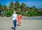 Adorable boy having fun with his mom on the tropical beach. White t-shirt, dark trousers and sunglasses. Barefoot on white sand.