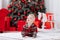 Adorable caucasian baby boy is sitting and holding gift toy Santa Claus near a Christmas tree, many festive gift boxed