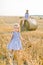 Adorable cute little baby girl in nice striped dress walking in summer wheat field. Pretty young mother sitting on hay