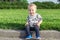 Adorable funny baby toddler boy smiling and sitting on road curb at city park with green grass on background. Cute little child