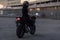 Adorable girl in black tight body suit and full-face helmet rides on stylish motorcycle at urban outdoors parking in